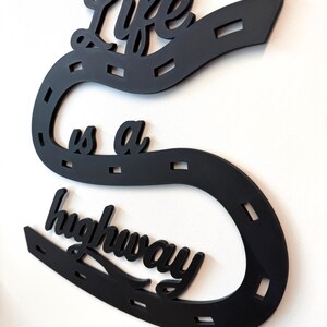 May include: A black wall-mounted sign shaped like a winding road with dashed white-style markings. The cutout text reads Life is a highway in a cursive script. The design features a continuous curved path that snakes across the wall surface.