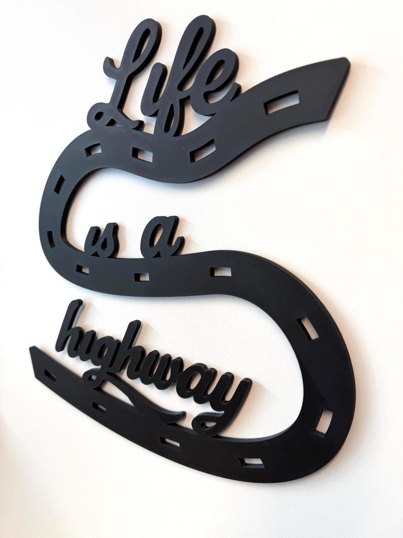 May include: A black wall-mounted sign shaped like a winding road with dashed white-style markings. The cutout text reads Life is a highway in a cursive script. The design features a continuous curved path that snakes across the wall surface.