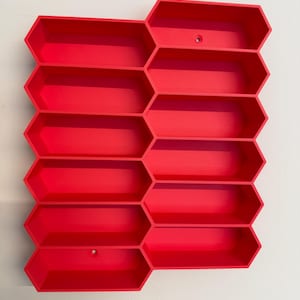 May include: A bright red wall-mounted storage unit featuring a modular honeycomb design with twelve elongated hexagonal compartments arranged in a staggered grid pattern, including two small circular mounting holes for installation.