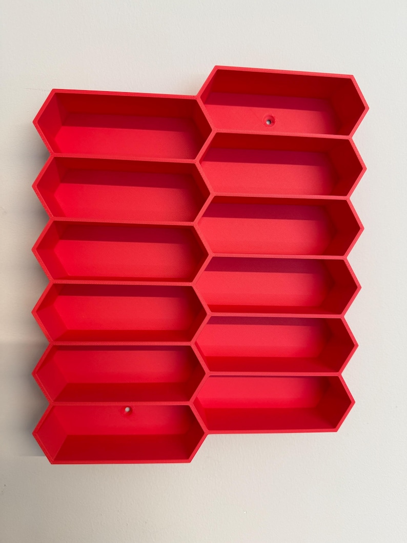 May include: A bright red wall-mounted storage unit featuring a modular honeycomb design with twelve elongated hexagonal compartments arranged in a staggered grid pattern, including two small circular mounting holes for installation.