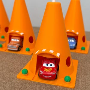 May include: Bright orange toy traffic cones with small toy cars inside. The cones have a white top and a small opening for the cars to enter. The cars are red, orange, and blue. Green decorative balls are at the base of the cones.