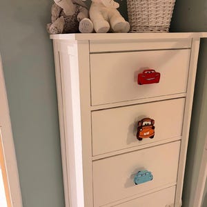 May include: White dresser with car-shaped drawer pulls in red, orange, and blue. The dresser has four drawers and is topped with stuffed animal toys. The wall behind the dresser is painted a light blue.