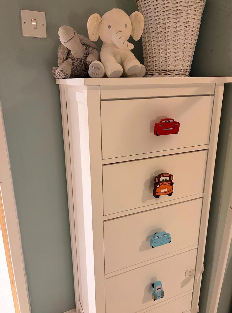 May include: White dresser with car-shaped drawer pulls in red, orange, and blue. The dresser has four drawers and is topped with stuffed animal toys. The wall behind the dresser is painted a light blue.