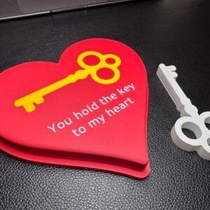 A red heart-shaped 3D-printed decoration with a gold key inset and the text ‘You hold the key to my heart.’ A silver key lies next to it. A perfect romantic Valentine’s gift.