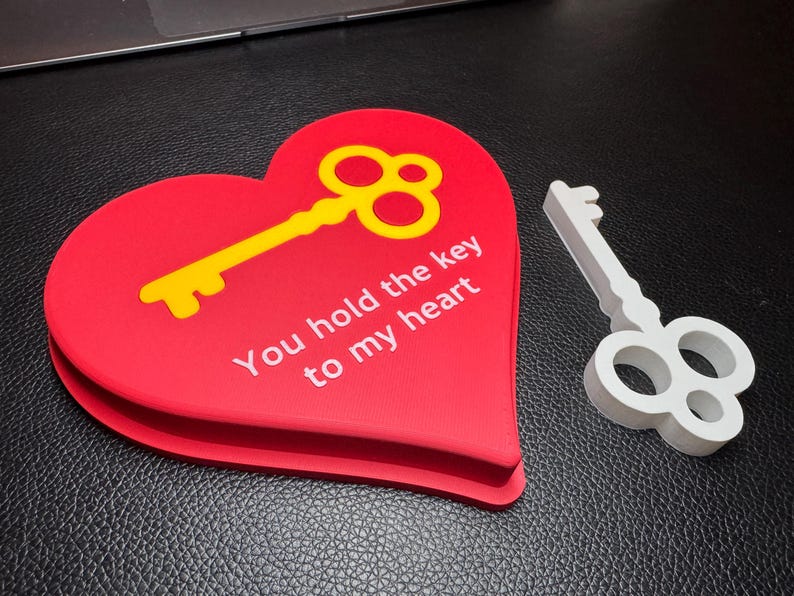 A red heart-shaped 3D-printed decoration with a gold key inset and the text ‘You hold the key to my heart.’ A silver key lies next to it. A perfect romantic Valentine’s gift.