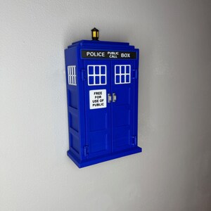 May include: A blue Police Public Call Box replica with white lettering and window details. The box has a small lamp on top and the text "FREE FOR USE OF PUBLIC" on the doors. The box is mounted on a white wall.