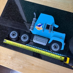 May include: A light blue toy semi-truck with black wheels and a silver exhaust stack. The truck has the word "DINOCO" printed on the side. A yellow and black tape measure is next to the truck.