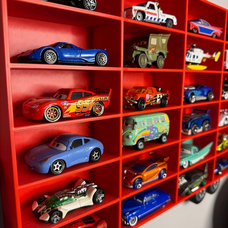 May include: A red display shelf filled with a collection of toy cars. The cars are of various colors and styles, including race cars, trucks, and vans. The shelf is made of red plastic and has multiple compartments to hold the cars.