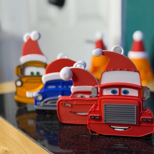 Może przedstawiać: Kolekcja kolorowych ozd&oacute;b samochodzik&oacute;w, z kt&oacute;rych każdy ozdobiony jest czapką Świętego Mikołaja. Samochody są w r&oacute;żnych kolorach, w tym czerwonym, niebieskim i ż&oacute;łtym. Ozdoby zaprojektowano z cechami kresk&oacute;wkowymi, idealne do dekoracji świątecznych.