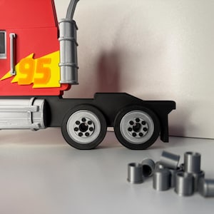 May include: A red toy semi-truck with the number "95" on the side in yellow and orange. The truck has black wheels and a silver exhaust stack. Several small, silver cylindrical pieces are in front of the truck.