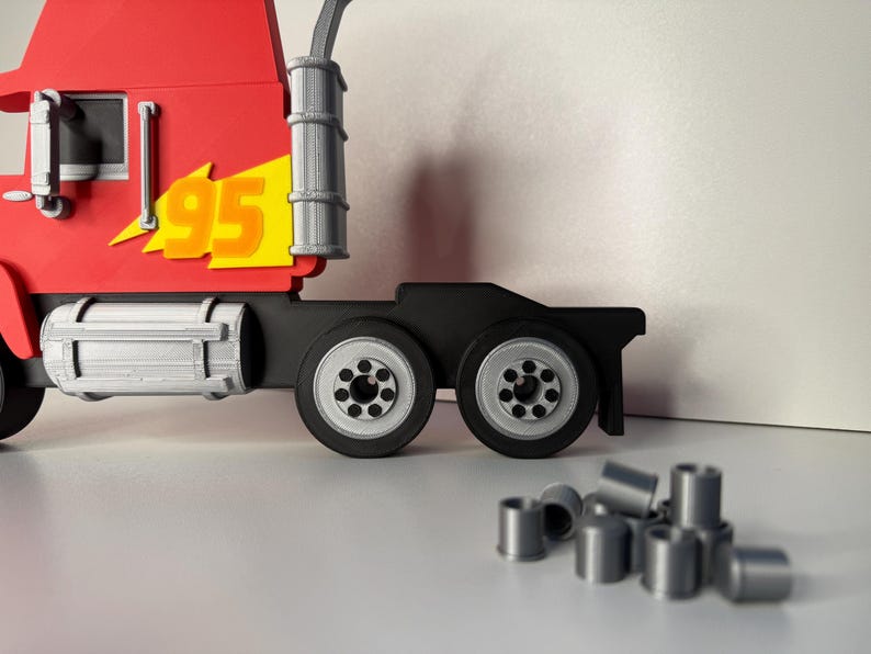 May include: A red toy semi-truck with the number "95" on the side in yellow and orange. The truck has black wheels and a silver exhaust stack. Several small, silver cylindrical pieces are in front of the truck.