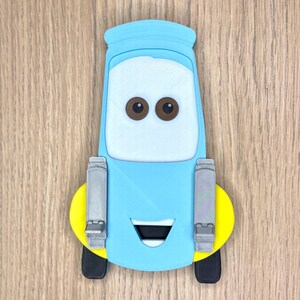 May include: A light blue cartoon car character with brown eyes, a white face, and a black mouth. The car has yellow and black wheels, and gray details. The character is a 3D printed item.