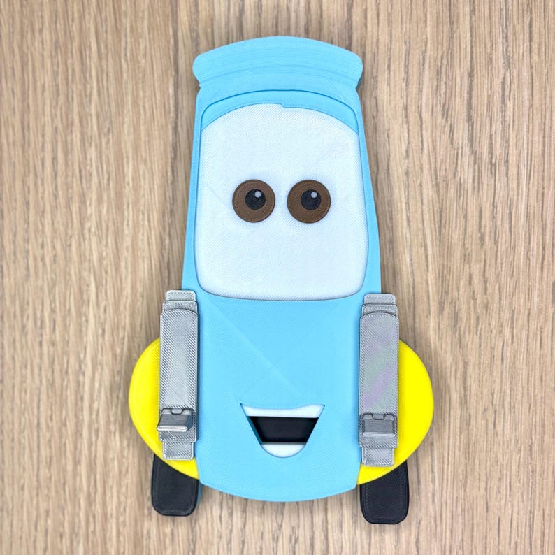 May include: A light blue cartoon car character with brown eyes, a white face, and a black mouth. The car has yellow and black wheels, and gray details. The character is a 3D printed item.