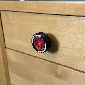 May include: A black and red drawer pull shaped like a tire, with the word "LIGHTYEAR" in white lettering. The drawer pull is mounted on a light-colored wooden cabinet. The cabinet has a second, silver-colored knob below the tire-shaped pull.