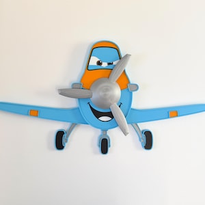 Könnte beinhalten: Eine Wanddekoration in Flugzeugform mit hellblauem und orangefarbenem Körper, einem lächelnden Gesicht, ausdrucksstarken Augen und einem markanten silbernen Dreiblattpropeller an der Vorderseite. Das Design umfasst kleine schwarze Räder und orange Akzente.