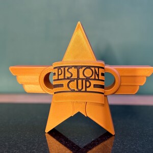 Puede incluir: Un trofeo dorado en forma de estrella con las palabras "PISTON CUP" en letras negras. El trofeo tiene extensiones en forma de alas y dos asas circulares. El dise&ntilde;o recuerda a un premio de carreras.