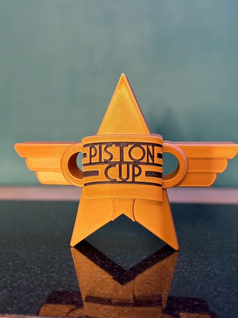 Puede incluir: Un trofeo dorado en forma de estrella con las palabras "PISTON CUP" en letras negras. El trofeo tiene extensiones en forma de alas y dos asas circulares. El dise&ntilde;o recuerda a un premio de carreras.