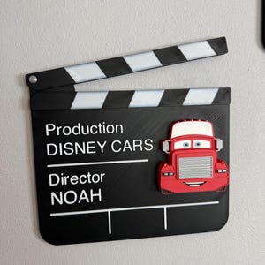 May include: A black film clapperboard with white stripes and text that reads "Production DISNEY CARS Director NOAH." A red cartoon truck is attached to the clapperboard.