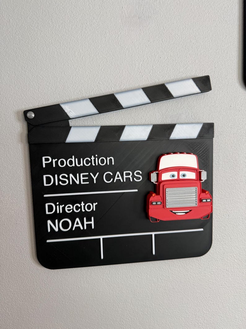 May include: A black film clapperboard with white stripes and text that reads "Production DISNEY CARS Director NOAH." A red cartoon truck is attached to the clapperboard.