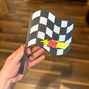 May include: A black and white checkered flag with a gray pole. The flag has a yellow lightning bolt with the number 95 in red. The flag is a racing theme.