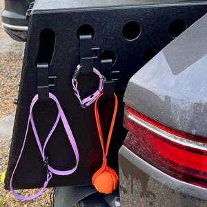 3 3D-printed hooks attached to the exterior of a DT dog crate, holding a purple lead, a matching collar, and an orange ball on a strap. The design keeps pet accessories tidy and easily accessible for outdoor use.