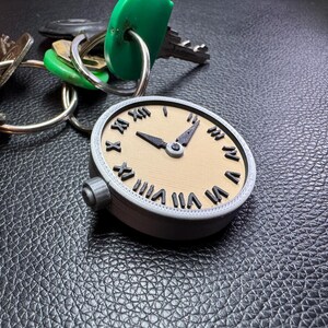 May include: A silver-coloured pocket watch with Roman numeral markings on a beige face. The watch is attached to a set of keys with green and silver keychains.