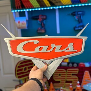 May include: A 3D-printed sign featuring the word "Cars" in silver lettering on a red and silver emblem. The sign has a triangular base and wing-like extensions. The sign is held by a person.