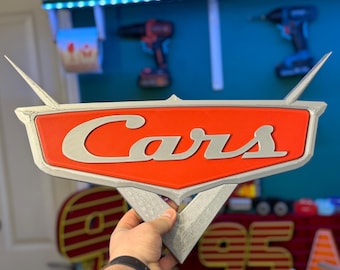 May include: A 3D-printed sign featuring the word "Cars" in silver lettering on a red and silver emblem. The sign has a triangular base and wing-like extensions. The sign is held by a person.