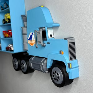 May include: A light blue toy truck with a trailer, mounted on a wall. The truck features black tires, a gray grill, and the "Dinoco" logo. The trailer has shelves holding miniature toy cars.