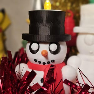 Adorable 3D-printed snowman tealight holders in white and multicolour, featuring festive designs with hats, scarves, and LED tealights. Perfect as Christmas decorations or gifts, displayed on a table with holiday-themed backdrops.