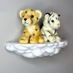 May include: Two plush animal toys, a cheetah and a white tiger, sit on a white cloud-shaped shelf. The cheetah is tan with black spots, while the tiger has black stripes. The shelf is mounted on a gray wall.
