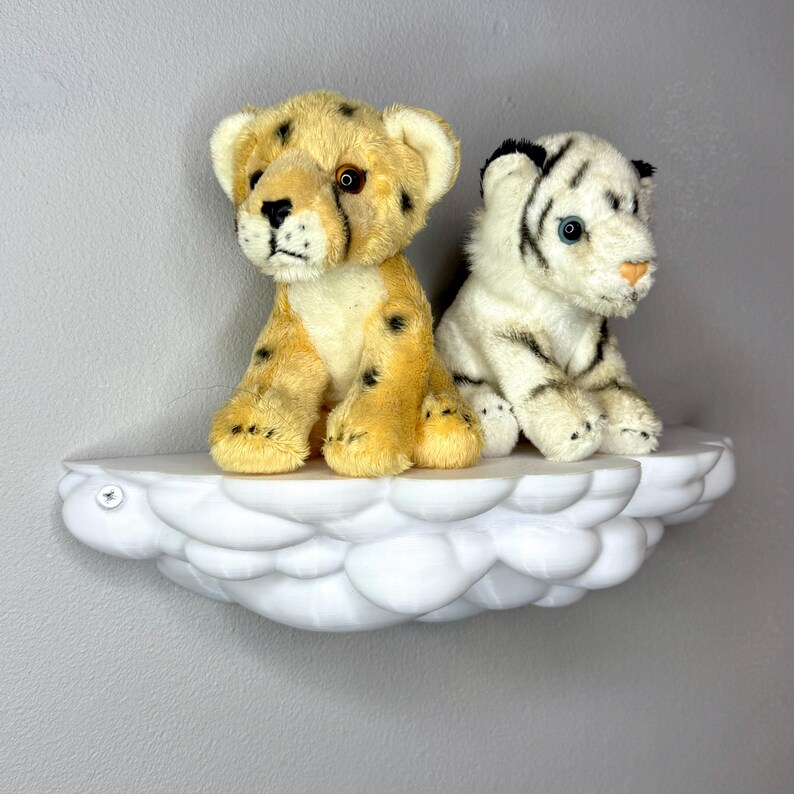 May include: Two plush animal toys, a cheetah and a white tiger, sit on a white cloud-shaped shelf. The cheetah is tan with black spots, while the tiger has black stripes. The shelf is mounted on a gray wall.
