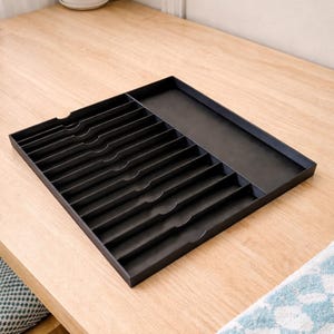 May include: A black rectangular cutlery tray with multiple compartments and a separate section. The tray is made of a dark material and sits on a light-colored wooden surface. A small plant in a white pot is visible in the background.