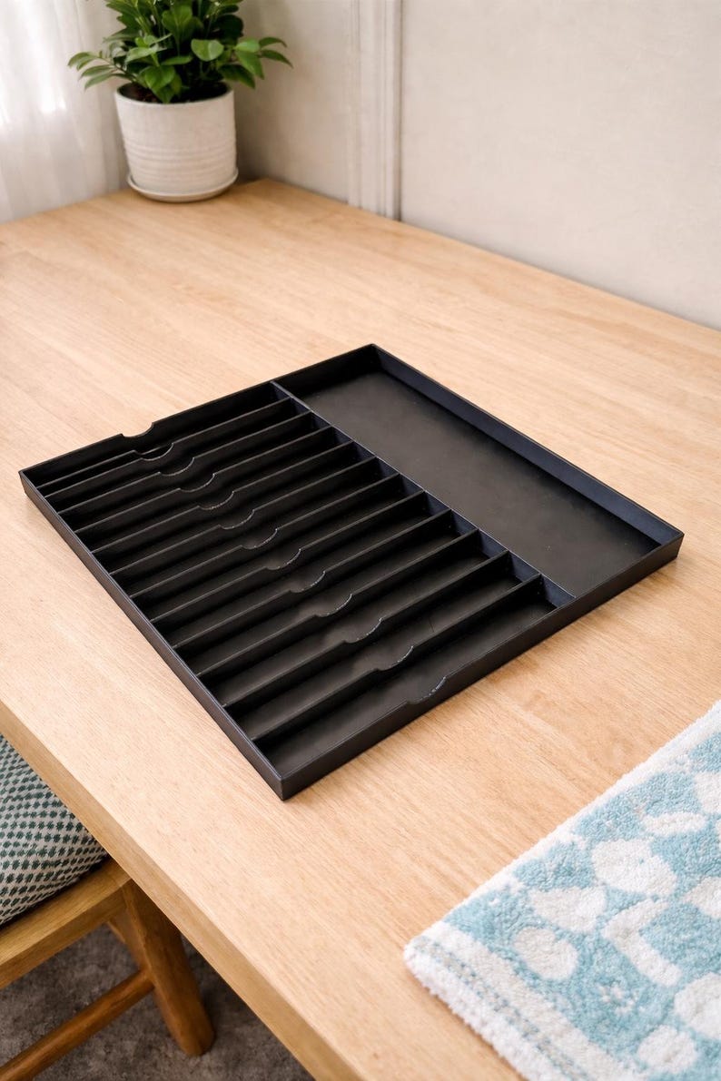 May include: A black rectangular cutlery tray with multiple compartments and a separate section. The tray is made of a dark material and sits on a light-colored wooden surface. A small plant in a white pot is visible in the background.