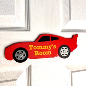 May include: A red car-shaped door sign with the text "Tommy's Room" in yellow. The sign has black wheels and a black window. The sign is mounted on a white door.