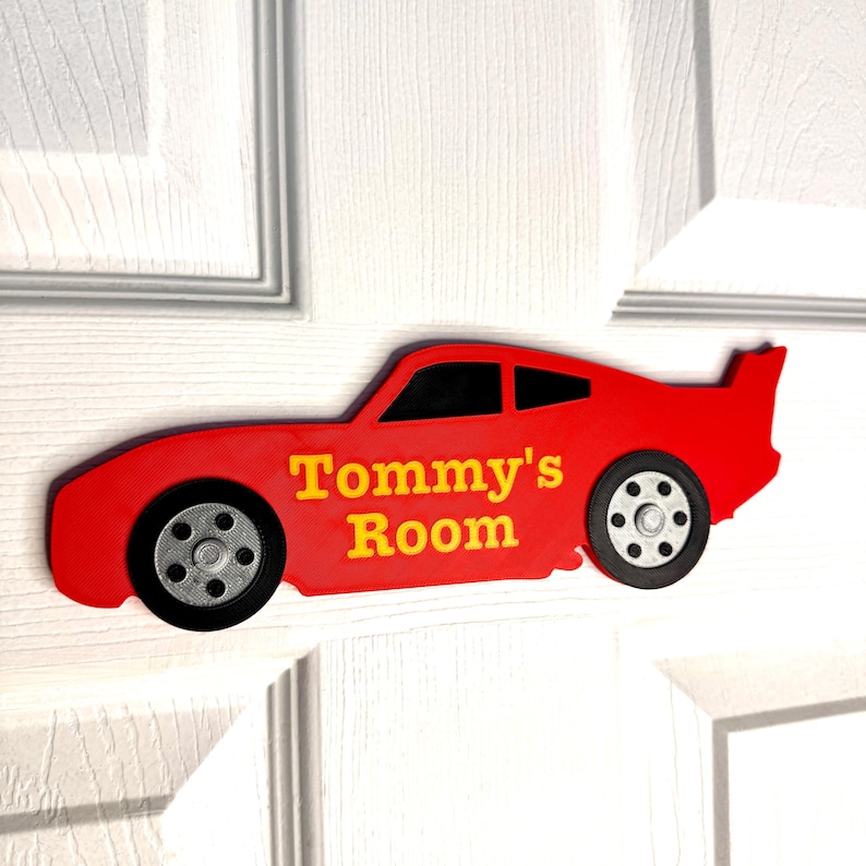 May include: A red car-shaped door sign with the text "Tommy's Room" in yellow. The sign has black wheels and a black window. The sign is mounted on a white door.