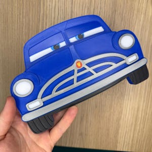 May include: A blue 3D-printed car-shaped object with a silver grille, white headlights, and blue eyes with white pupils. The car features a small red and yellow circular emblem on the front grille and black tires at the bottom corners.