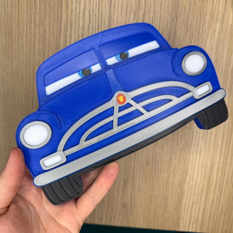 May include: A blue 3D-printed car-shaped object with a silver grille, white headlights, and blue eyes with white pupils. The car features a small red and yellow circular emblem on the front grille and black tires at the bottom corners.