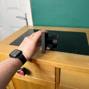 以下が含まれることがあります： 正方形のベースと円筒形のハンドルが付いた黒いキャビネットノブ。ノブは、黒いカウンタートップが付いた木製のキャビネットの前に保持されています。黒い時計が人の手首にあります。