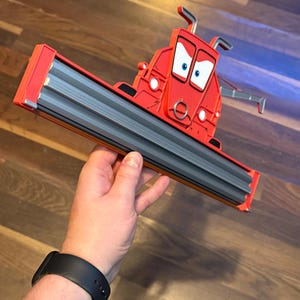 May include: A red plastic character figure resembling a combine harvester with a wide grey front header attachment. The figure features a cartoon face with large white eyes, blue pupils, and a silver ring detail on the front. The structure is angular.