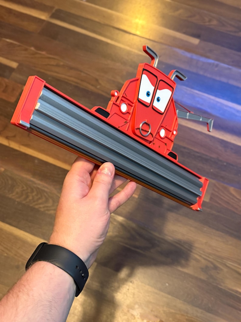 May include: A red plastic character figure resembling a combine harvester with a wide grey front header attachment. The figure features a cartoon face with large white eyes, blue pupils, and a silver ring detail on the front. The structure is angular.
