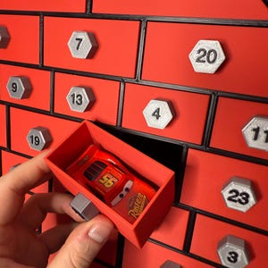 May include: A red advent calendar with numbered silver hexagonal knobs. A red toy car is in a red drawer. The calendar has a black border and is set against a white background.