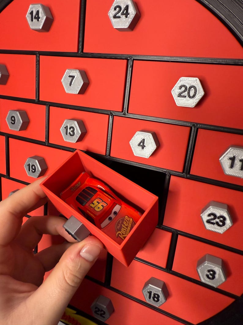 May include: A red advent calendar with numbered silver hexagonal knobs. A red toy car is in a red drawer. The calendar has a black border and is set against a white background.