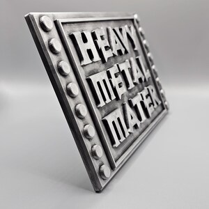 Peut inclure: Panneau rectangulaire argent&eacute; avec les mots "HEAVY METAL MATER" en lettres en relief. Le panneau a une bordure rivet&eacute;e avec des accents circulaires. Le panneau est inclin&eacute;, mettant en valeur son design en trois dimensions.