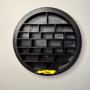 May include: A black, circular wall shelf made from a tire, featuring multiple rectangular compartments. A yellow lightning bolt with the word "Bowie" is attached to the bottom. The shelf is mounted on a white wall.