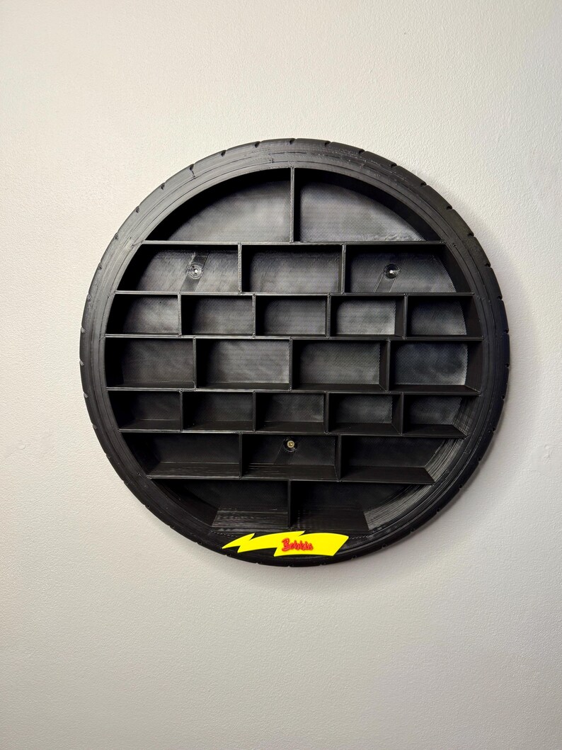 May include: A black, circular wall shelf made from a tire, featuring multiple rectangular compartments. A yellow lightning bolt with the word "Bowie" is attached to the bottom. The shelf is mounted on a white wall.