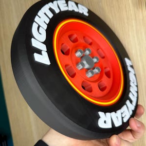 May include: A 3D-printed black tire with a red rim and yellow accent. The tire features the white text "LIGHTYEAR" around the outer edge. The wheel has a silver hub with five lug nuts. The item is held in a person's hand.