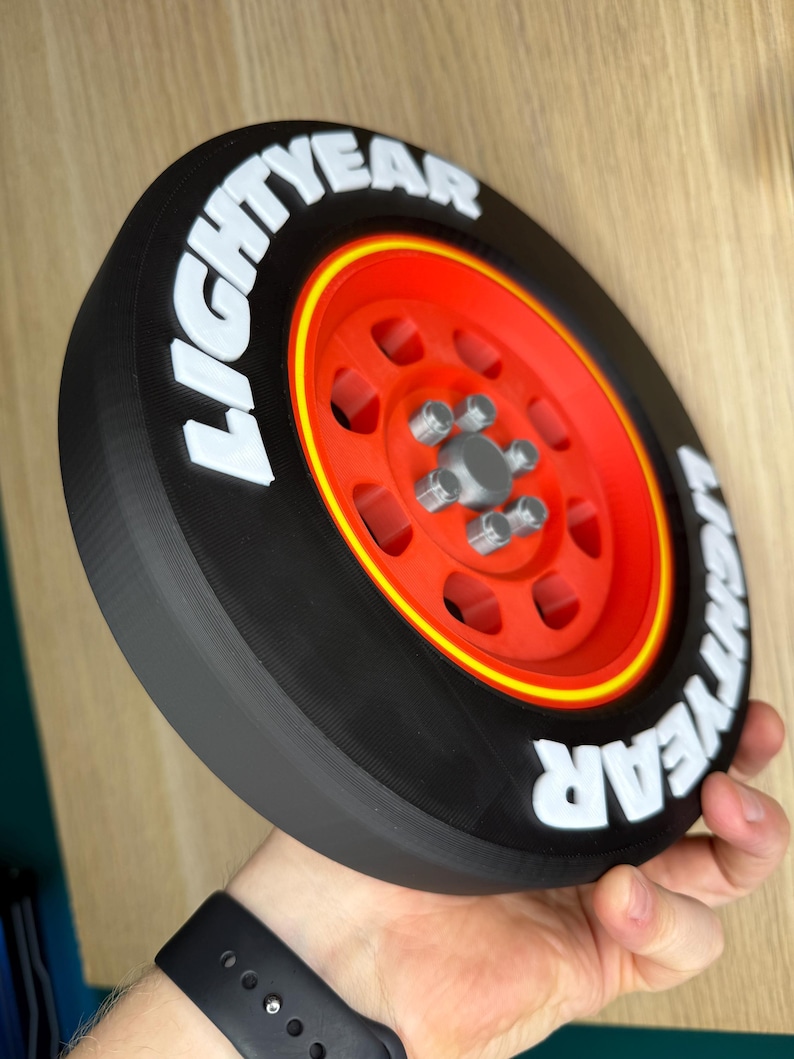 May include: A 3D-printed black tire with a red rim and yellow accent. The tire features the white text "LIGHTYEAR" around the outer edge. The wheel has a silver hub with five lug nuts. The item is held in a person's hand.
