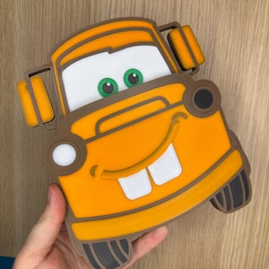 May include: An orange and brown cartoon car character with a smiling face, white teeth, and green eyes. The car has a rectangular shape with rounded edges and two rectangular shapes on the top. The car is held by a hand.