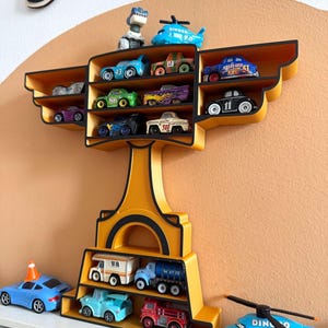 Puede incluir: Un estante de exhibici&oacute;n amarillo y negro con forma de trofeo, lleno de coches de juguete y un helic&oacute;ptero. El estante tiene m&uacute;ltiples compartimentos para guardar los veh&iacute;culos de juguete. El fondo es una pared de color naranja claro.